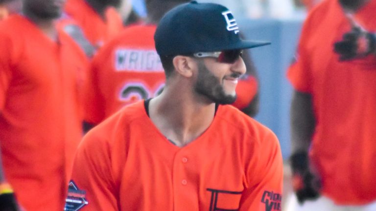 Colin Kaepernick smiling in an orange jersey and dark cap at a celebrity softball game