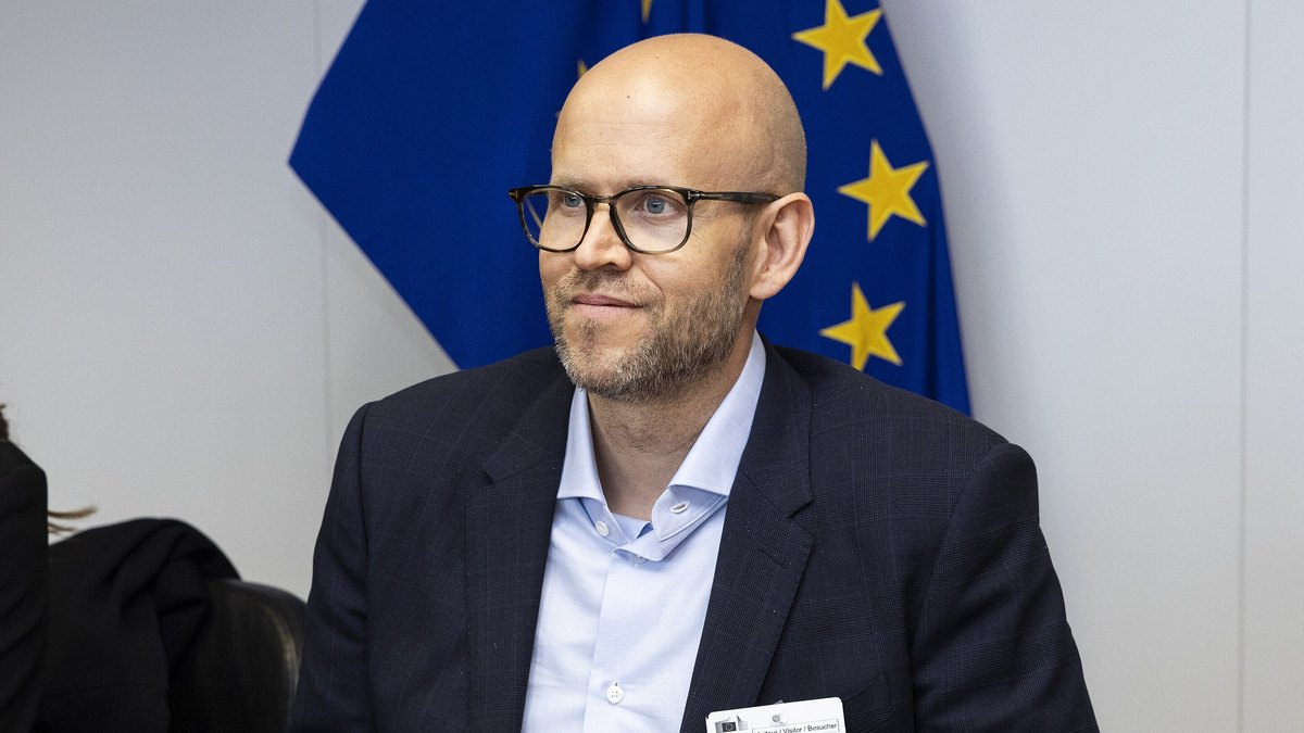 Daniel Ek in glasses and a dark blazer seated at a meeting table in front of an EU flag