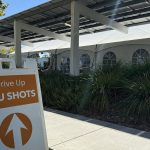 A sidewalk sign reading Drive Up Flu Shots with an arrow, in front of a white tent under a solar canopy