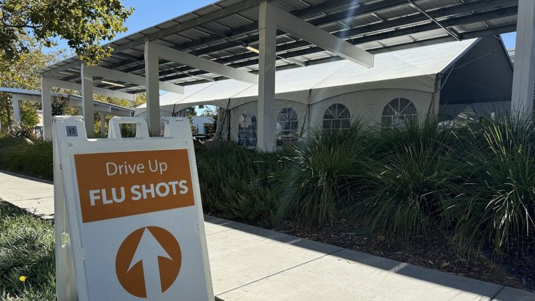 A sidewalk sign reading Drive Up Flu Shots with an arrow, in front of a white tent under a solar canopy