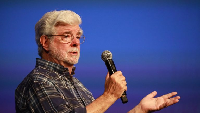 George Lucas, with white hair, glasses and a plaid shirt, speaking into a microphone against a blue background