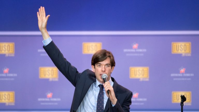 John Mulaney in a dark suit holding a microphone and raising one hand on stage in front of a USO backdrop