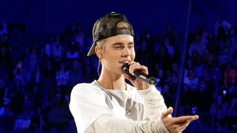Justin Bieber singing into a microphone on stage in a white shirt and backward cap