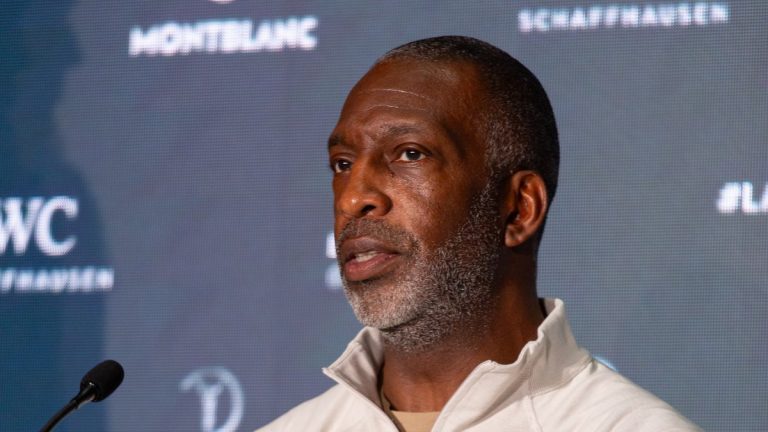 Michael Johnson, in a white zip-neck top, speaking at a press conference in front of a sponsor backdrop
