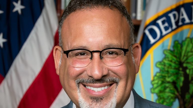 Miguel Cardona smiling in front of a U.S. flag and the Department of Education seal