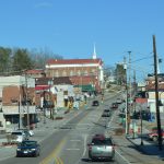 Morgan Avenue (U.S. Route 58 Alternate) running uphill through downtown Pennington Gap, with brick storefronts, a pharmacy sign and a church on the hill under a clear blue sky