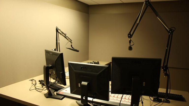 Empty radio studio with microphones on boom arms above computer monitors and a mixing console