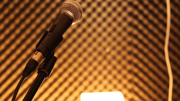 Close-up of a handheld vocal microphone on a stand in warm light against an acoustic foam wall