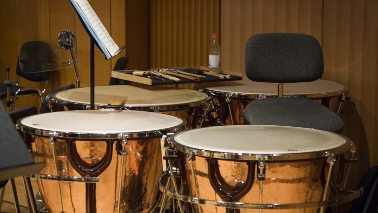 A set of copper orchestral timpani with mallets on a rack in a wood-paneled rehearsal room