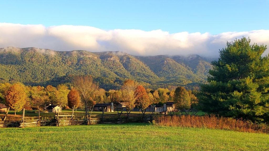 Fields, split-rail fence and log buildings at Wilderness Road State Park below a forested mountain ridge on a fall morning