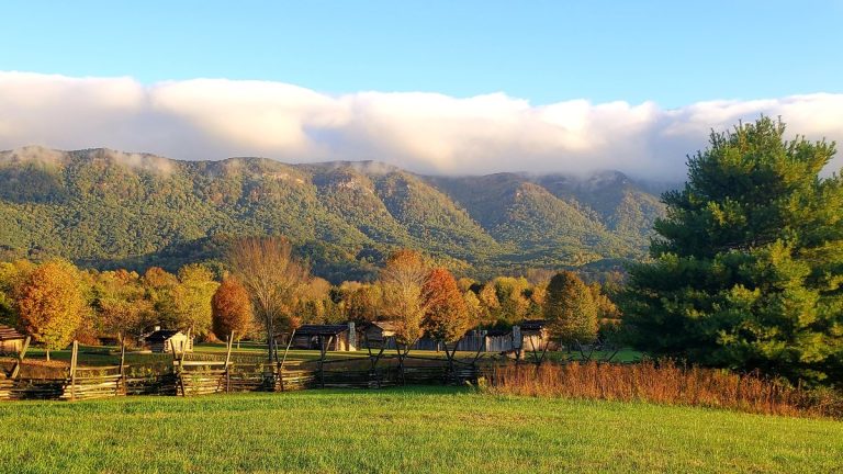 Fields, split-rail fence and log buildings at Wilderness Road State Park below a forested mountain ridge on a fall morning